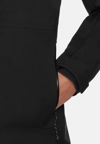 Mano parzialmente all'interno della tasca laterale di un giubbotto nero con zip e dettagli con fibbia sulla manica.