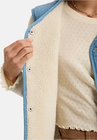 Spijkervest met witte teddy voering, drukknopen en zachte textuur. Daaronder een crème top met patroon en gegolfde randen.