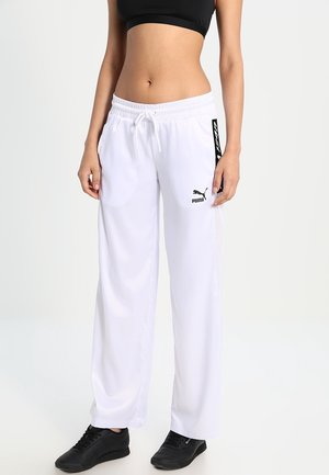 Femme portant un pantalon de survêtement blanc Puma, un crop top noir et des baskets noires, debout devant un fond clair uni.