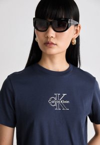 Navy cotton t-shirt featuring white embroidered "Calvin Klein" logo. Model wears black cat-eye sunglasses and large gold hoop earrings.
