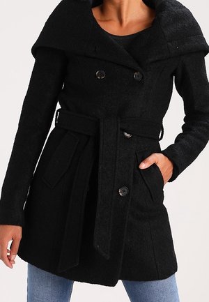 Femme portant un manteau noir croisé ceinturé avec un large col, une main dans la poche, associé à un jean bleu.