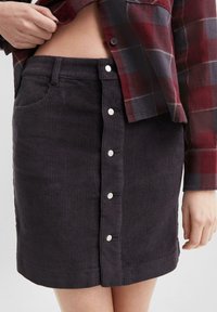 Donkergrijze corduroy rok met verticale ribbels, een getailleerde vorm, een voorzak en zilveren knopen in het midden voor sluiting.
