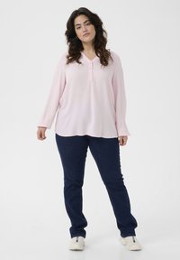 Blusa de manga larga rosa claro con un acabado texturizado y un escote en V que presenta tres botones, combinada con pantalones de mezclilla azul oscuro y zapatillas blancas.