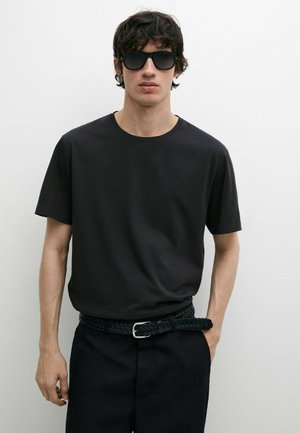 Jonge man met zwarte zonnebril, zwart T-shirt, gevlochten zwarte riem en zwarte broek tegen een effen witte achtergrond.