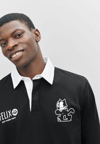 Polo negro con cuello blanco, que presenta un diseño bordado de Felix el Gato y texto en el lado izquierdo. Tejido suave, corte casual.
