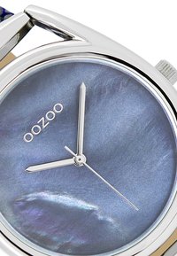 Montre-bracelet argentée avec un cadran rond, un cadran en nacre bleue, des aiguilles des heures et des minutes en argent, et le nom de la marque "oozoo" affiché.