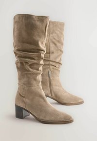 Beige suède kniehoge laarzen met een slouch-ontwerp, puntige neus en een houten blokhak. Voorzien van een rits aan de zijkant voor gemakkelijke instap.
