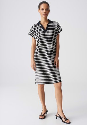 Donna in piedi, indossa un vestito a righe orizzontali bianche e nere fino al ginocchio con colletto nero e sandali neri con tacco e cinturini.