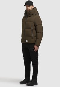 Uomo che indossa una giacca imbottita verde oliva con cappuccio, pantaloni cargo neri, stivali neri e un cappellino nero, in piedi contro uno sfondo neutro.
