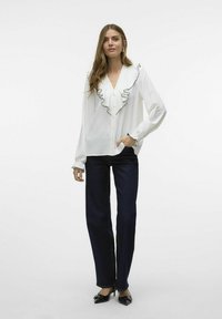 Blusa blanca con tejido texturizado y ribete de volantes negro en el cuello; combinada con jeans oscuros de pierna ancha y zapatos negros de tacón bajo.