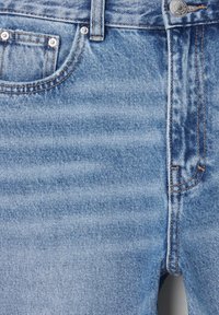 Gros plan sur l'avant d'un jean en denim bleu clair avec un bouton en métal, des passants pour ceinture, une poche avant et des détails de couture de couleur cuivre.