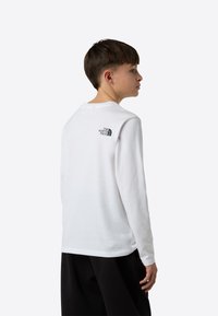 Hosszú ujjú fehér ing, pamut anyagból, kerek nyakkivágással, a hátán fekete logóval, sötét nadrággal viselve.