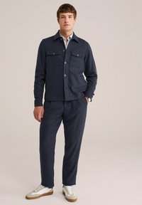 Marineblauwe wolmix overhemdjas met twee borstzakken, gecombineerd met bijpassende losse broek en witte sneaker met beige accenten.