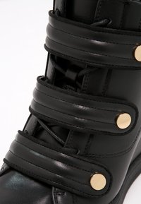 Svart läderkänga med flera texturerade remmar, guldhardwaredetaljer och en rundad tådesign. Slät yta med synliga snörningar.