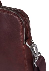 Bolso de cuero marrón con cremalleras plateadas, de textura suave, bordes redondeados y un cierre metálico. Cuenta con una correa de tela ajustable.