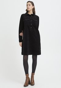 Rochie cămașă din catifea neagră cu nasturi în față, buzunare și mâneci lungi. Purtați cu colanți negri și ghete maro până la gleznă.