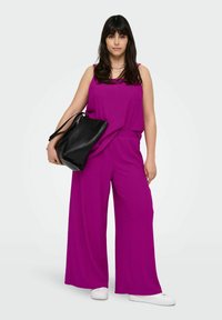 Salopette fuchsia avec des jambes larges et une coupe décontractée, présentant une texture côtelée. Associée à un sac noir et des baskets blanches.