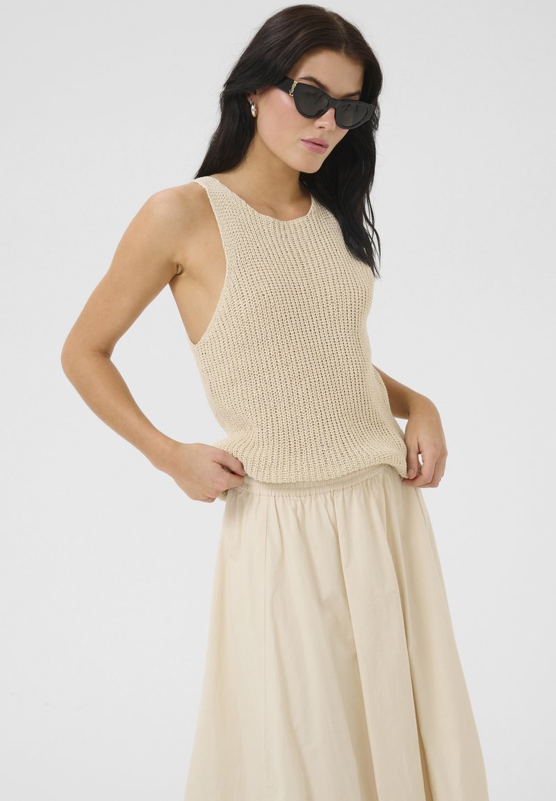 Virkat beige linne med öppna sidor, matchat med en svävande beige kjol. Modellen håller kjolen för att visa dess mjukhet och fall. Svarta solglasögon kompletterar stilen.