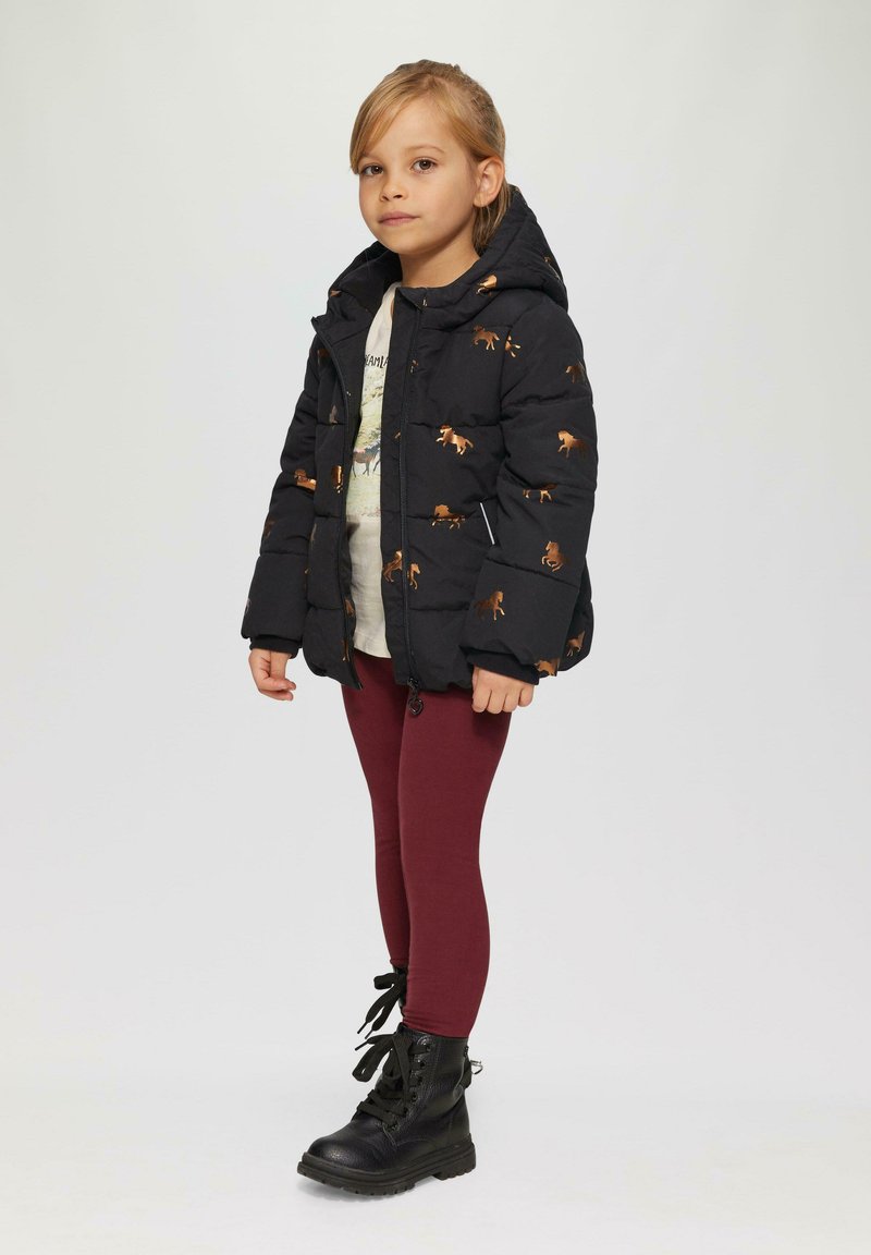 Doudoune noire avec des motifs animaliers bruns, fermeture éclair, poches latérales, portée sur une chemise claire, associée à des leggings bordeaux et des bottes noires.