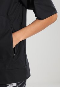 Černý sportovní top s krátkými rukávy, texturovaná tkanina, boční zipová kapsa a volný střih. Stylizováno s vzorovanými legínami pro kontrast.