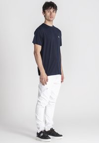 Mladý muž s krátkymi tmavými vlasmi, oblečený v tmavomodrom tričku, bielych roztrhnutých džínsoch a čiernych teniskách, stojí pred jednoduchým bielym pozadím.