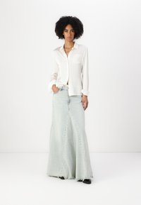 Camisa blanca de botones con un bolsillo en el pecho, combinada con una falda larga de mezclilla azul clara que presenta bordes deshilachados y detalles en las costuras laterales.