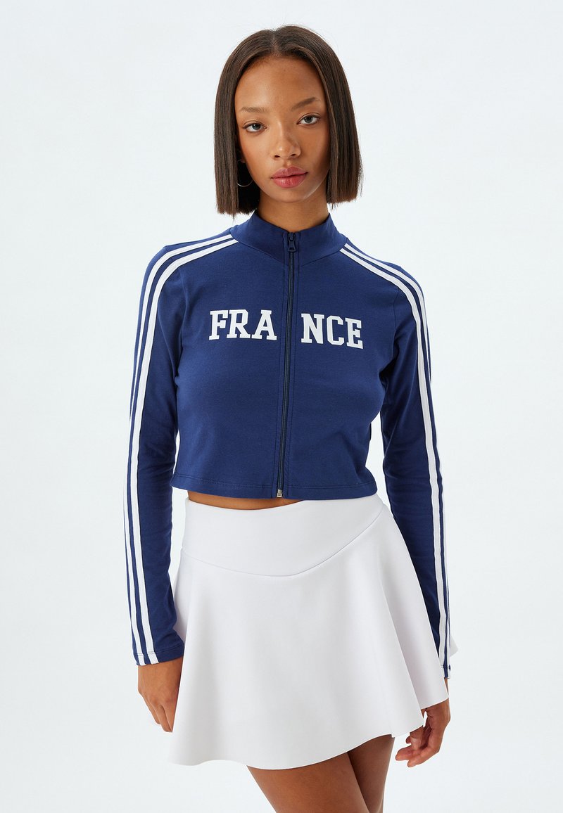 Chaqueta de cremallera azul marino con el texto "FRANCE" en blanco, con rayas horizontales blancas a lo largo de las mangas. Acompañada de una falda blanca acampanada.