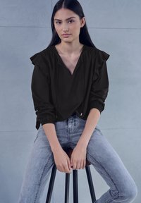 Blouse noire avec épaules à volants, col en V et détails à cordon, associée à un jean skinny gris clair. Texture de tissu lisse.