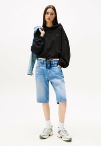 Sudadera negra con capucha, corte holgado, pantalones cortos de mezclilla de lavado azul claro, calcetines blancos y zapatillas grises. Chaqueta colgada sobre el hombro.