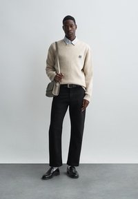 Bežs vilnas džemperis ar V veida kaklu un mazu logotipu, uzvilkts virs gaišas svītrainas krekliņa. Melni bikses un ādas kurpes noslēdz izskatu.