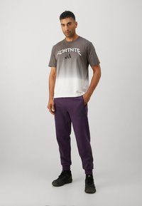 Szara koszulka z gradientem z napisem "FORTNITE" i logo adidas, w połączeniu z ciemnofioletowymi spodniami dresowymi. Obie części odzieży mają luźny krój.