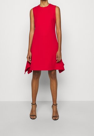 Röd ärmlös klänning med rund halsringning, A-linjeformad skärning och asymmetrisk fåll. Tillverkad av mjukt tyg, kombinerad med svarta gladiatorsandaler med klack.