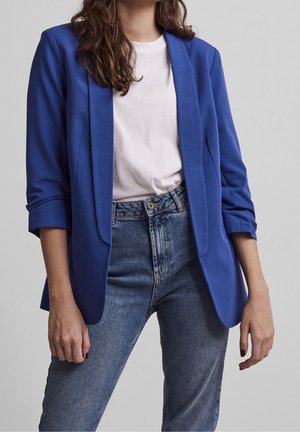 Frau trägt einen königsblauen Blazer über einem weißen T-Shirt und blaue hoch taillierte Jeans vor einem schlichten grauen Hintergrund.