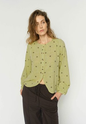 Donna con capelli ondulati indossa una camicetta verde chiaro con bottoni e piccoli motivi floreali scuri, abbinata a pantaloni scuri a righe, con le mani nelle tasche.