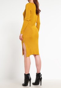 Femme portant une robe tricotée à manches longues jaune moutarde avec une fente sur le côté, associée à des bottines noires à talons hauts, se tenant de côté.