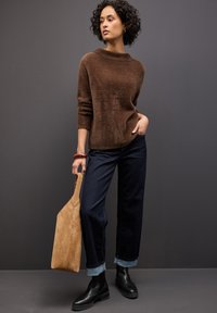 Brauner, flauschiger Pullover mit hohem Kragen, blaue, weite Jeans mit umgeschlagenen Säumen und schwarze Stiefeletten. Trägt eine beigefarbene Wildleder-Tasche.