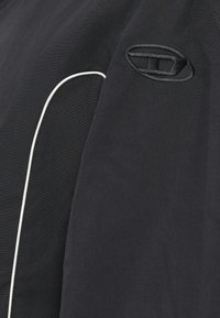 Μαύρο μπουφάν με αντίθετες λευκές ραφές. Διαθέτει υφασμάτινη υφή και κεντημένο λογότυπο στον αριστερό ώμο.