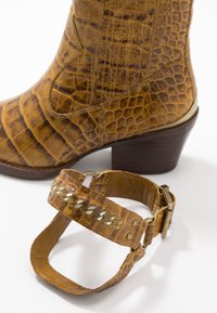 Bota de cuero grabado en cocodrilo marrón con punta cuadrada y tacón apilado, combinada con una correa marrón a juego que presenta detalles metálicos.