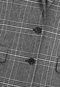 Tissu à motif pied-de-poule noir et blanc avec deux boutons ronds noirs cousus et une poche partiellement visible.