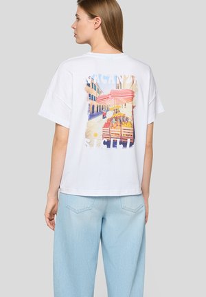 Frau mit glatten braunen Haaren, die ein weißes T-Shirt mit buntem Street-Markt- und rotem Regenschirm-Print trägt, kombiniert mit hellblauen Jeans.