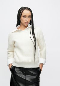 Valkoinen neulottu pusero, jossa on teksturoitu kuvio, pyöreä pääntie ja käännetyt hihansuut, yhdistetty mustaan nahkahameeseen.