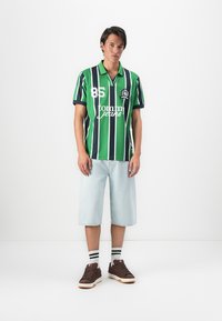 Young man wearing green and navy striped polo, light blue denim shorts, striped socks, and brown sneakers standing against white background.