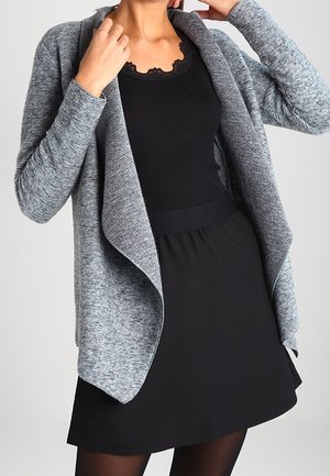 Femme portant une robe noire avec un décolleté festonné et un cardigan gris ouvert à l'avant, posant avec les bras levés près de sa tête.