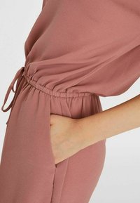 Oblečení v prachově růžové barvě s elastickým pasem a šňůrkou. Volný střih s bočními kapsami. Měkká, texturovaná látka.