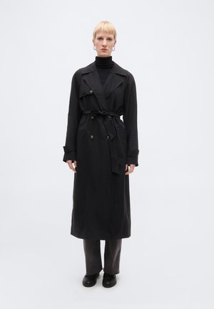 Person iført en lang sort trenchcoat bundet i taljen over en sort top og mørke bukser, stående mod en ensfarvet hvid baggrund.