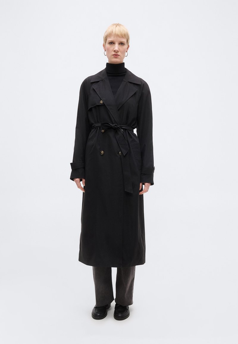 Άτομο που φορά μακρύ μαύρο παλτό τρανς, δεμένο στη μέση, πάνω από μαύρο μπλουζάκι και σκούρο παντελόνι, που στέκεται μπροστά σε λευκό φόντο.