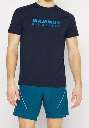 Mann trägt ein dunkelblaues T-Shirt mit dem Text "Mammut Since 1862" und türkisfarbene Sportshorts mit hellblauen Akzenten, steht vor einem weißen Hintergrund.