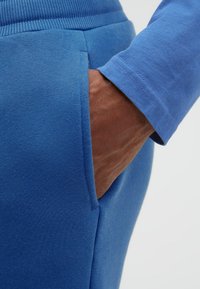Hand in der Tasche, trägt einen blauen Pullover und passende blaue Jogginghosen, Nahaufnahme zeigt Stofftextur und Nähte im Detail.