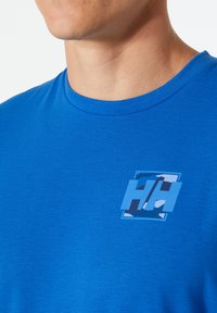 Modrá tkaninová trička s kulatým výstřihem. Obsahuje potištěné modro-bílé logo na hrudi v obdélníkovém tvaru.