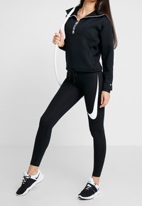Zwarte sporttrui met een ritskraag, gecombineerd met zwarte leggings met een grote witte Nike swoosh. Zwarte sportschoenen maken de outfit compleet.